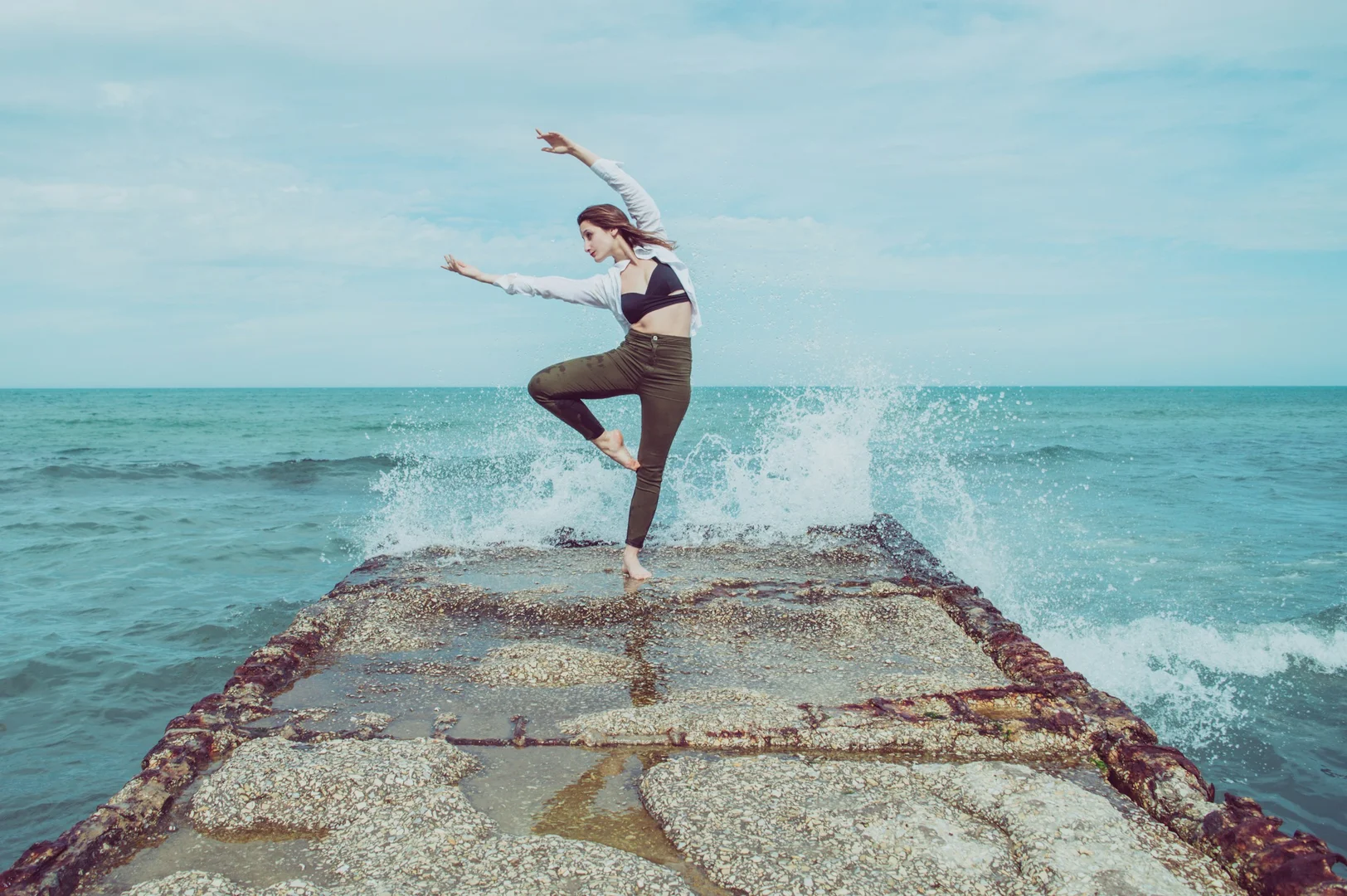 A dancer balanced on a sea-splashed jetty as waves break behind her, illustrative of Aszure Barton's earthy, texture-driven choreographic style