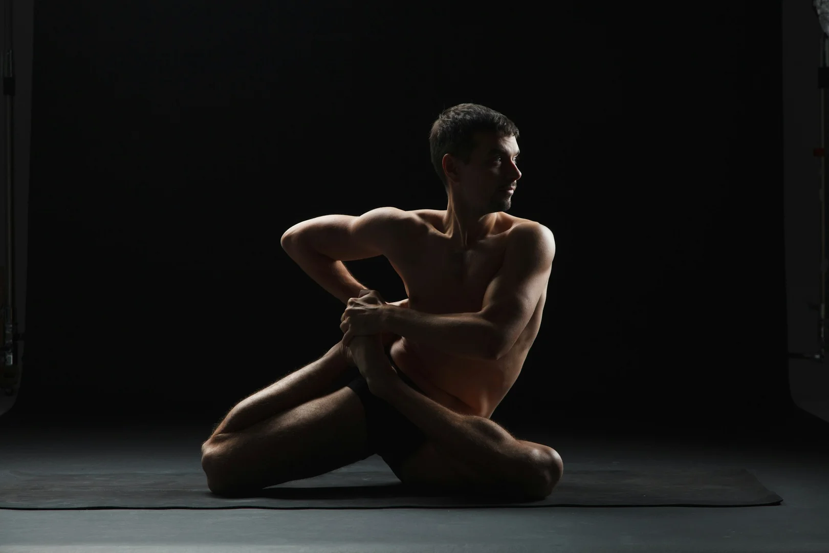 A dancer in a tightly coiled, contorted seated pose against a dark backdrop, illustrative of Wayne McGregor's cognitively driven physical vocabulary