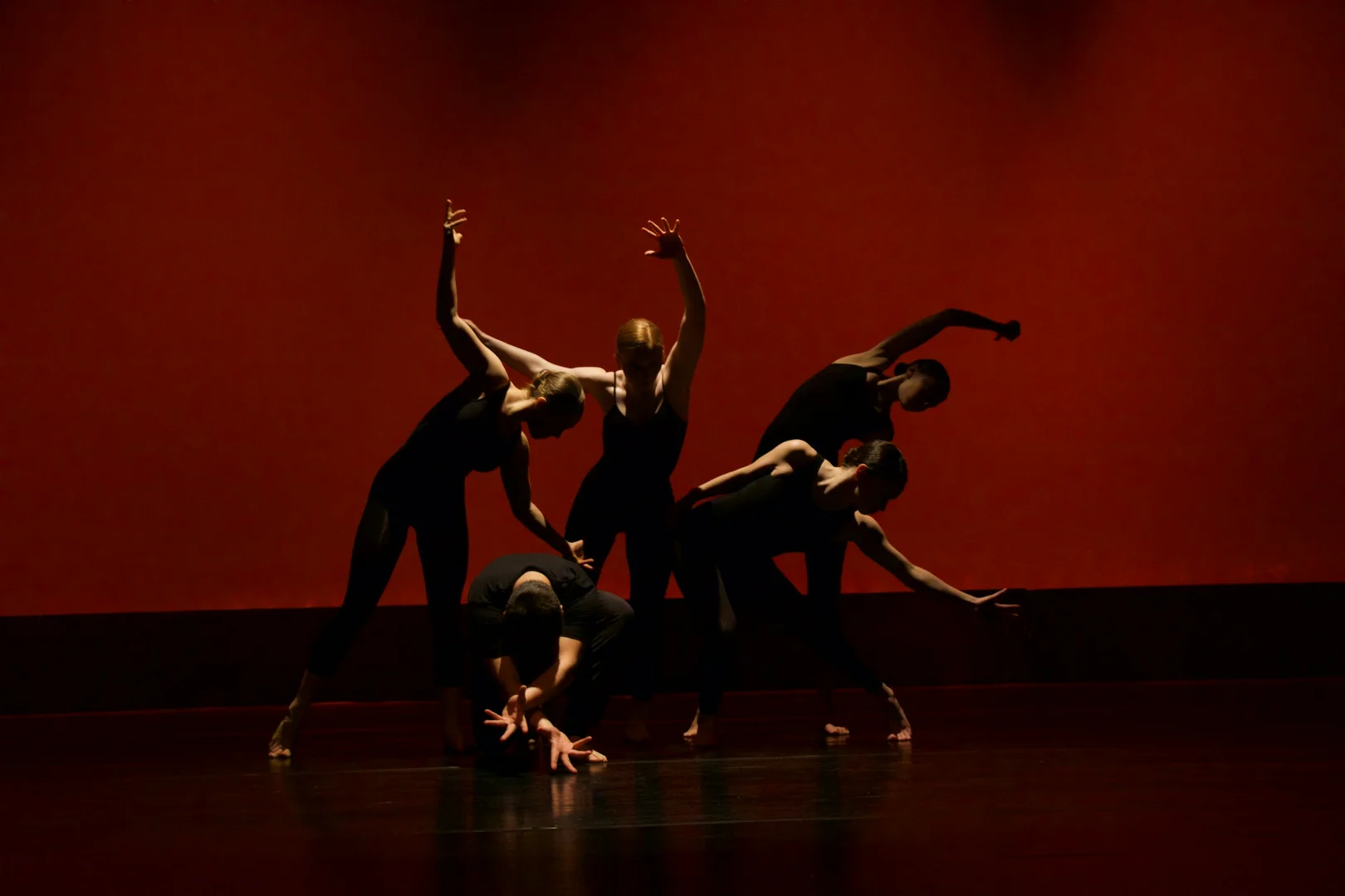 An ensemble of dancers in sharp, grounded shapes under a deep red wash, illustrative of the sensation-driven Gaga movement language